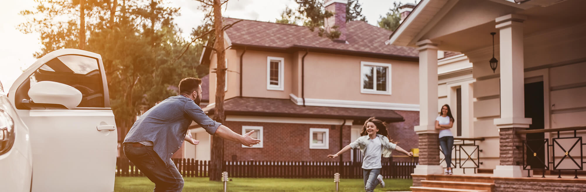 Young father coming home from work, daughter running across the yard to create him and mother watching from the front porch